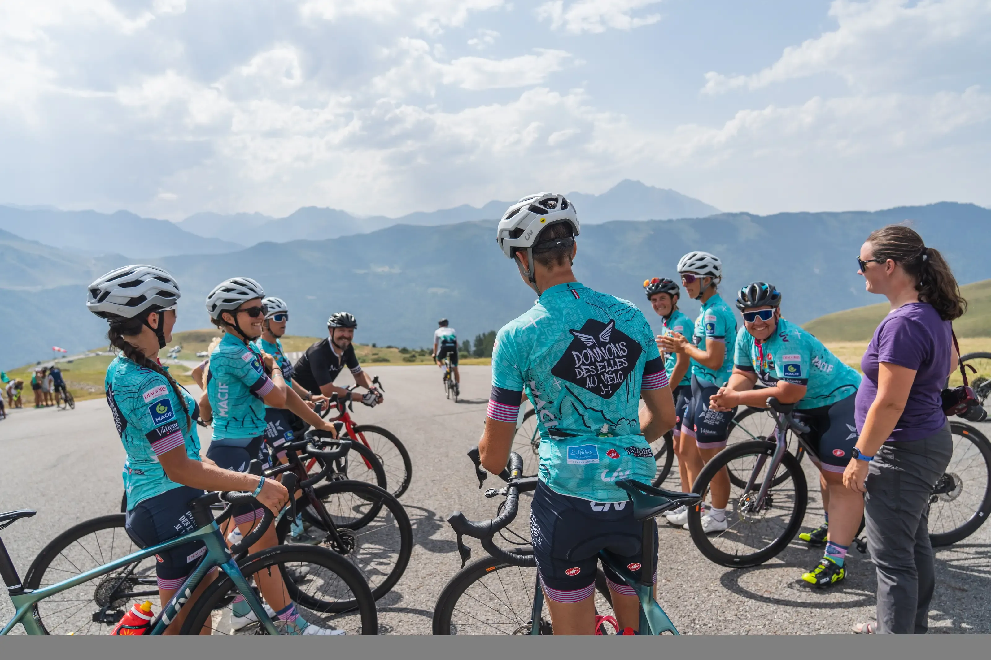 Un groupe de cyclistes portant des maillots turquoise fait une pause avec leurs vélos sur une route de montagne, discutant chaleureusement face à un panorama de sommets en arrière-plan.